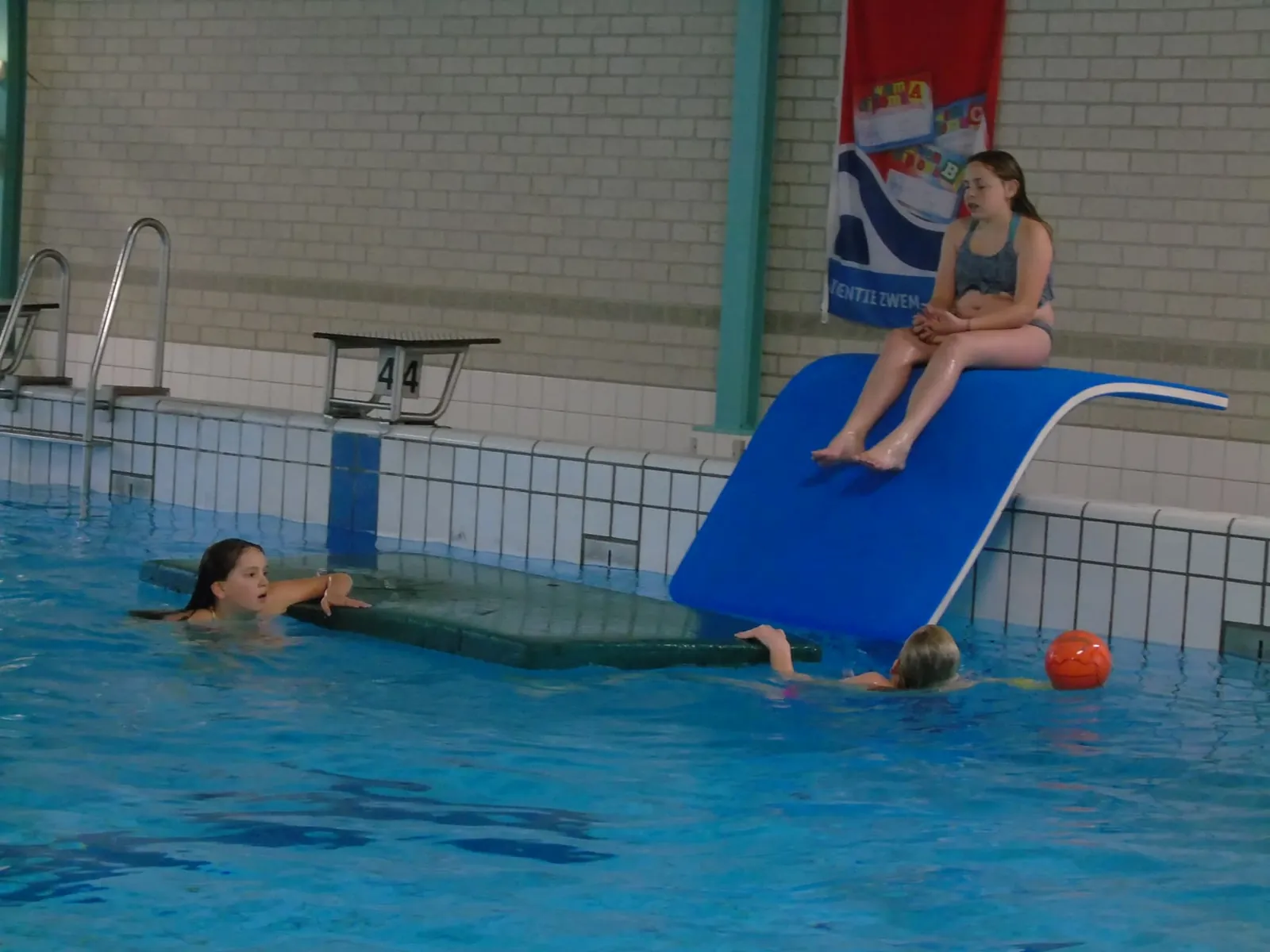 Meisje zit bovenaan een blauwe glijmat aan de badrand, terwijl twee kinderen bij een drijvende mat en een bal in het water zwemmen