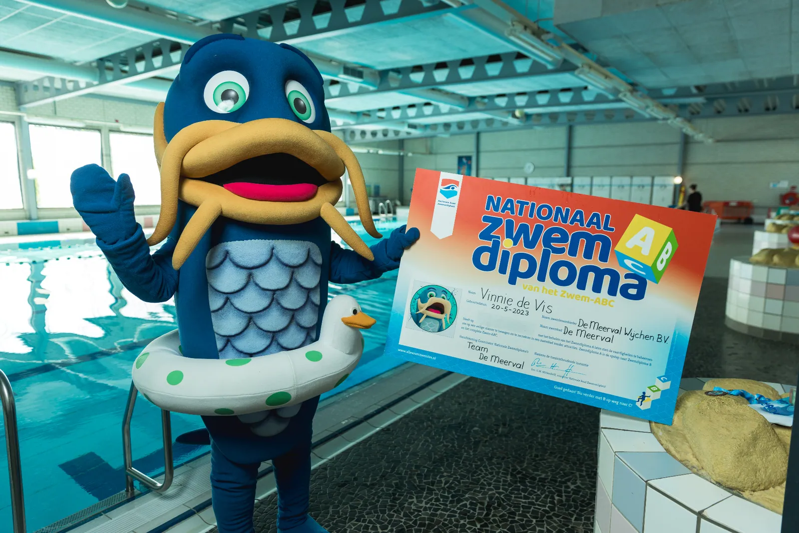 Vinnie de Vis, de mascotte van Zwembad De Meerval, toont trots een Nationaal Zwemdiploma van het Zwem-ABC bij het zwembad