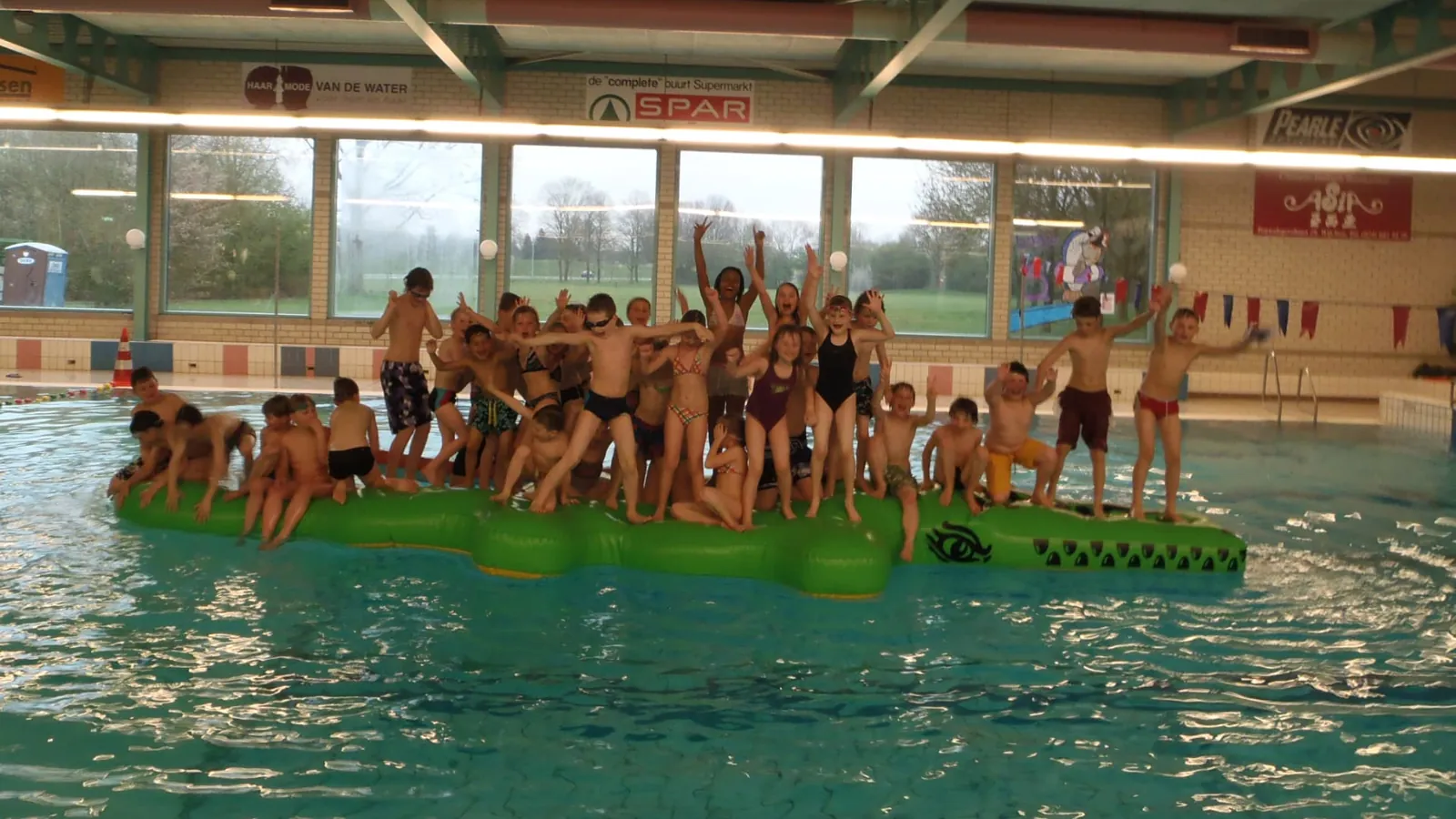 Grote groep kinderen staat juichend met de armen omhoog op een groene opblaaskrokodil in het zwembad